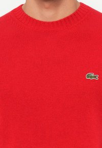 Pulover tricotat roșu cu guler rotund, având un mic logo verde de crocodil pe pieptul stâng. Textură moale și design ajustat.