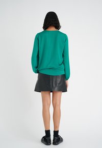 Blusa verde a maniche lunghe con scollatura rotonda, abbinata a una mini gonna di pelle nera. Il modello indossa calze nere e sandali.