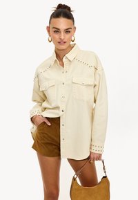 Camisa de botón color crema con acentos de tachuelas en los hombros, que presenta dos bolsillos frontales y mangas largas. Combinada con pantalones cortos marrones.