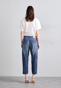 Lehký bílý bavlněný tričko v kombinaci s modrými jeansy s mírným vzestupem a volným střihem, které mají cargo kapsy a hnědou nášivku na zadní straně.