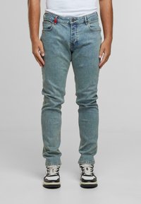 Muškarac obučen u svijetloplave slim-fit traperice i bijele i crne tenisice, stoji naslonjen na jednostavnu svijetlosivu pozadinu.