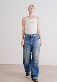 Witte tanktop met "BOSS" logo, gecombineerd met lichtblauwe oversized denim jeans met een beschadigde knie. Groene schoenen maken de outfit compleet.