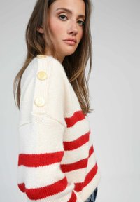 Pull en tricot crème avec des rayures rouges, comportant des boutons aux épaules et un coupe ample et texturé. Tissu doux avec un motif de tricot visible.