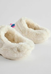 Chaussons en fausse fourrure couleur crème avec une texture douce, présentant un bout rond et un accent coloré décoratif sur le dessus.