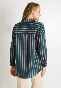 Camisa a rayas verdes con rayas verticales rosas y blancas, mangas largas, puños con botones y dobladillo redondeado. La tela parece ligera y texturizada.