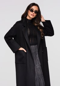 Manteau noir surdimensionné avec un col à revers, deux poches avant et des manches amples, porté sur un haut noir transparent et une jupe grise plissée.