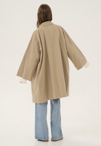 Béžový oversized kabát s límcem, širokými rukávy a žebrovanými pletenými manžetami, nošený přes světle modré rozšířené džíny. Hladká textilie.