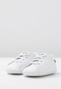 White leather sneakers with gray accents, perforated details, and green branding on the heel. Rounded toe and laced closure. Flat rubber sole.