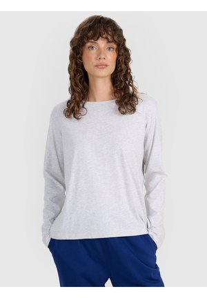 Mujer con cabello rizado y marrón que lleva una camiseta gris claro de manga larga y pantalones azules, de pie con las manos en los bolsillos frente a un fondo liso.
