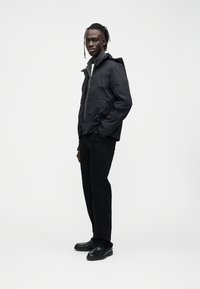 Juoda vandeniui atspari striukė su gobtuvu, priekyje užtrauktuku ir šoninėmis kišenėmis; derinama su juodomis kelnėmis ir batais. Lygus audinys, minimalistinis dizainas.