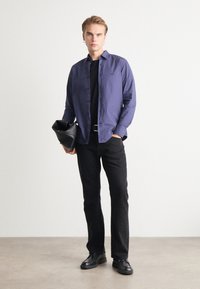 Cămașă cu mânecă lungă, cu nasturi, de culoare albastru închis, asortată cu un tricou negru, pantaloni negri și pantofi negri. Modelul ține o geantă neagră.