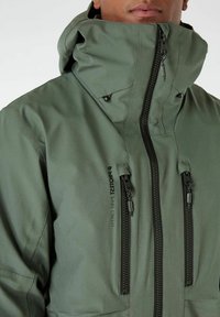 Giacca impermeabile verde con colletto alto, dotata di due tasche con zip, zip nere e texture opaca. Logo stampato sul fronte.