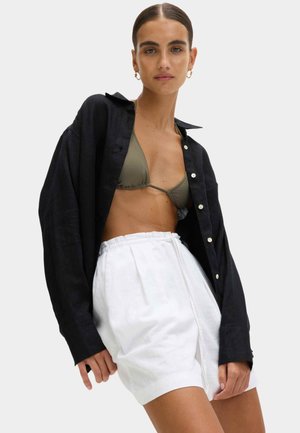 Chemise en lin noire avec détails de boutons portée sur un haut de bikini vert olive et un short blanc à taille élastique, mettant en valeur les textures et les couleurs.