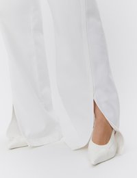 Picioare purtând pantaloni albi cu croială largă și fante laterale, asortați cu pantofi albi cu toc ascuțit pe fundal alb.