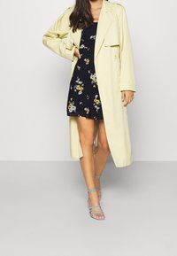 Trench coat amarillo claro sobre un vestido floral azul marino de mangas cortas. Presenta tacones azules con tiras. Tejido suave y corte relajado.
