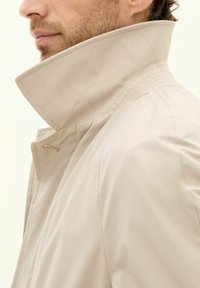 Giacca beige con colletto alto, caratterizzata da un motivo a diamante e dettagli cuciti. Il materiale sembra liscio e leggero.