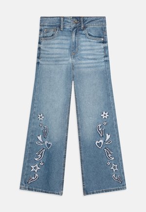 Jean en denim bleu avec jambes larges et motifs brodés blancs représentant des fleurs, des cœurs et des étoiles sur les deux jambes du pantalon.