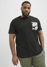 T-shirt noir à manches courtes avec une poche à motifs présentant des designs en coup de pinceau blancs et gris, porté avec un pantalon vert clair.