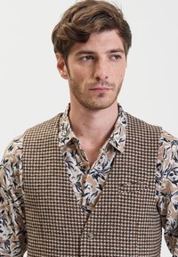 Gilet a scacchi marroni sopra una camicia floreale beige. Il gilet presenta un rever a punta e bottoni frontali, mescolando trame e motivi.