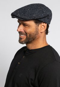 Cappello piatto in herringbone blu navy realizzato in tessuto testurizzato, con una corona arrotondata e una visiera corta e rigida. Abbinato a una maglietta henley nera.