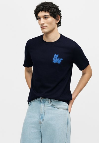 Άνδρας με σγουρά μαλλιά, που φοράει μπλε σκούρο t-shirt με ένα μικρό μπλε κουνέλι και ανοιχτό μπλε τζιν, τα χέρια πίσω από την πλάτη.