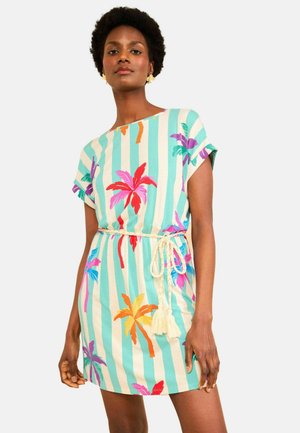 Žena oblečená v krátkom rukáve, šaty po kolená s tyrkysovými a bielymi vertikálnymi pruhmi a farebnými potlačami palmových stromov, previazaná béžovým lanovým opaskom.