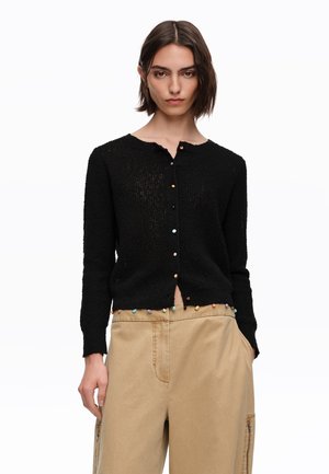 Joven mujer con un cárdigan negro abotonado con botones redondos multicolores y pantalones anchos beige, de pie frente a un fondo blanco.