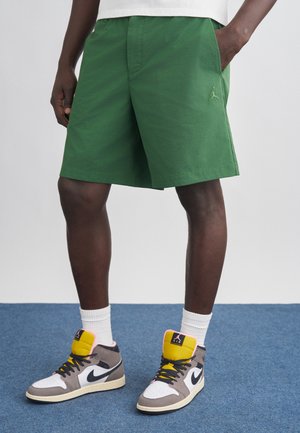 Mies, jolla on vihreät shortsit, valkoiset sukat ja moniväriset Nike Air Jordan -korkeavartiset tennarit, seisoo sinisellä matolla.