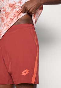 Κόκκινα αθλητικά σορτς από ελαφρύ ύφασμα, με πορτοκαλί λωρίδα στο πλάι και μικρό λογότυπο στο κάτω αριστερό πόδι. Ορατή ελαστική ζώνη στη μέση.