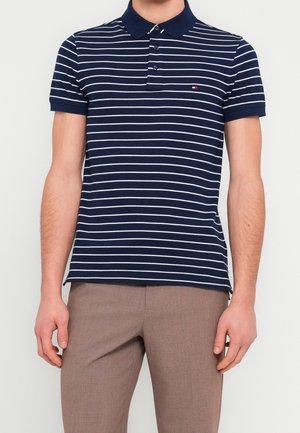 Homme portant un polo à manches courtes rayé bleu marine et blanc avec un pantalon marron, debout devant un fond clair uni.