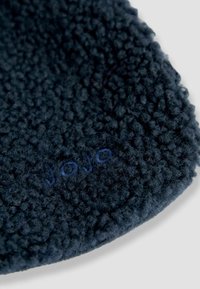 Tamsiai mėlyna flisinė skrybėlė su švelniu, tekstūriniu paviršiumi ir subtiliu mėlynu logotipu, siuvinėtu ant krašto. Apvalus, minimalistinis dizainas.