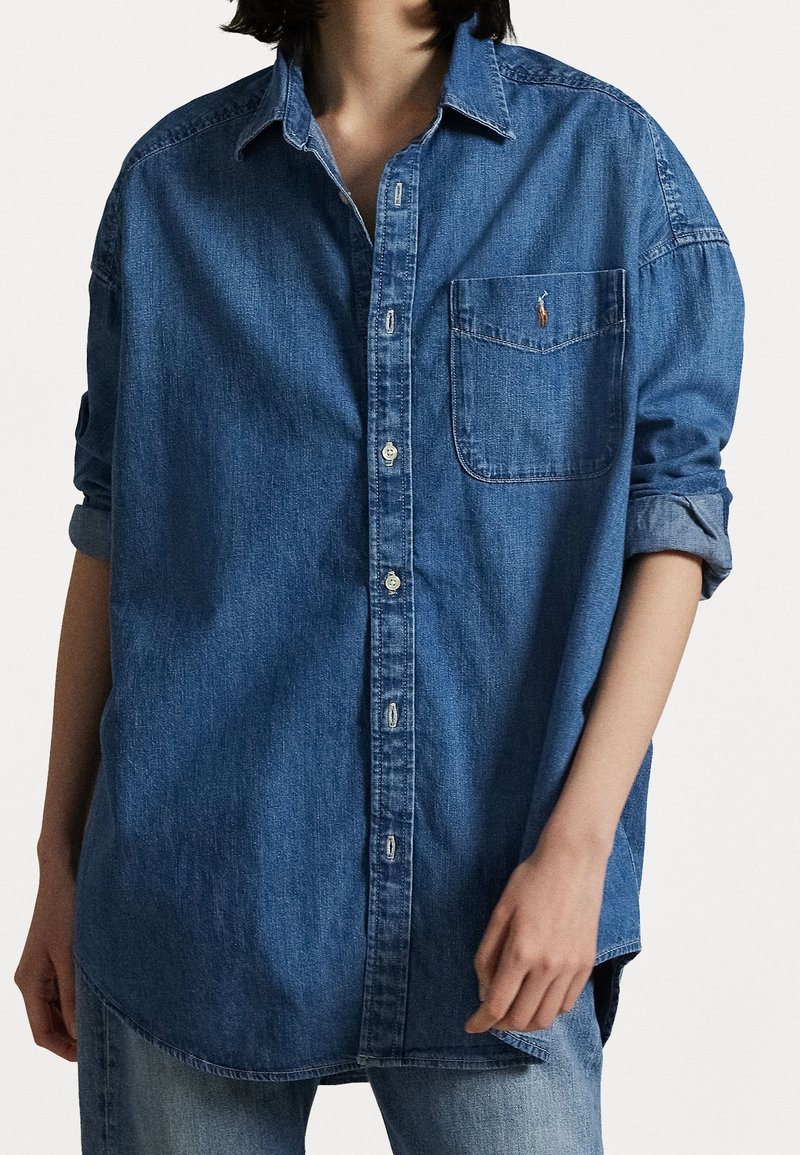 Dunkelblaue Denim-Hemdbluse mit Brusttasche, hochkrempelbaren Ärmeln und abgerundetem Saum. Der Stoff hat eine leichte Textur.
