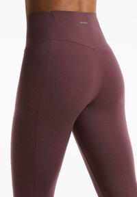 Közelkép egy személyről, aki magas derekú, testhezálló lila leggingset visel, rajta "OYSHO" logóval a derékpánton.