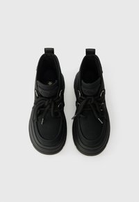 Μαύρα αθλητικά παπούτσια με στρογγυλή μύτη, υφασμάτινο επάνω μέρος από υλικό με υφή, χοντρή ελαστική σόλα και έντονες κορυφές δεσίματος. Διαθέτουν επενδεδυμένο κολάρο και λεπτομέρεια λογότυπου.