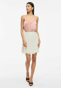 Camisole en satin rose clair avec des bordures en dentelle, associé à une jupe fluide crème. Le modèle porte des sandales à talons carrés noirs. Design simple et textures douces.