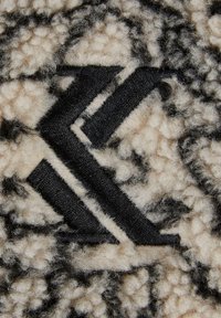 Teksturoitu, pehmeä beige kangas mustalla geometrisella logolla. Sellaisessa on pehmeä, pörröinen pinta ja tummempia kontrastiväriä.