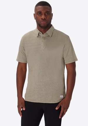 Uomo che indossa una polo manica corta verde oliva chiaro e pantaloni neri, in piedi di fronte a uno sfondo semplice.