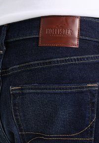 Tmavě modré džíny s hnědou koženou nášivkou s embosovaným nápisem "Hollister California". Obsahují zlaté švy a detaily na zadních kapsách.