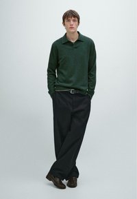 Ciemnozielony, textured sweter z kołnierzykiem, zestawiony z luźnymi czarnymi spodniami; model nosi ciemnobrązowe buty. Prosty, minimalistyczny styl.
