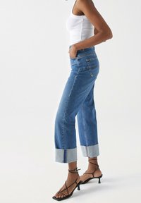 Jeans recortados de mezclilla azul con un puño de un azul claro, diseño de cintura alta y bolsillos laterales. Combinados con tacones negros con tiras.