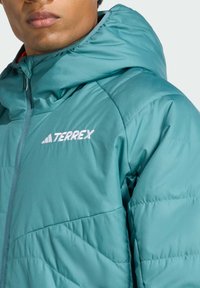 Türkise Daunenjacke mit Kapuze, gestepptem Textil und weißem "TERREX"-Logo. Mit einem Reißverschluss auf der Vorderseite und elastischen Bündchen. Leichtes Design.