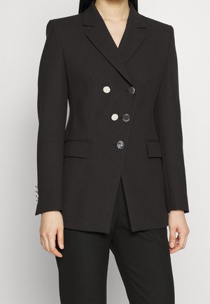 Femme portant un blazer noir cintré à double boutonnage avec des boutons argentés et un pantalon noir assorti, debout devant un fond uni.