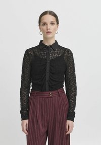 Blusa in pizzo nero con chiusura a bottoni, maniche lunghe e collo; caratterizzata da dettagli arricciati sul torso, abbinata a pantaloni a righe bordeaux.