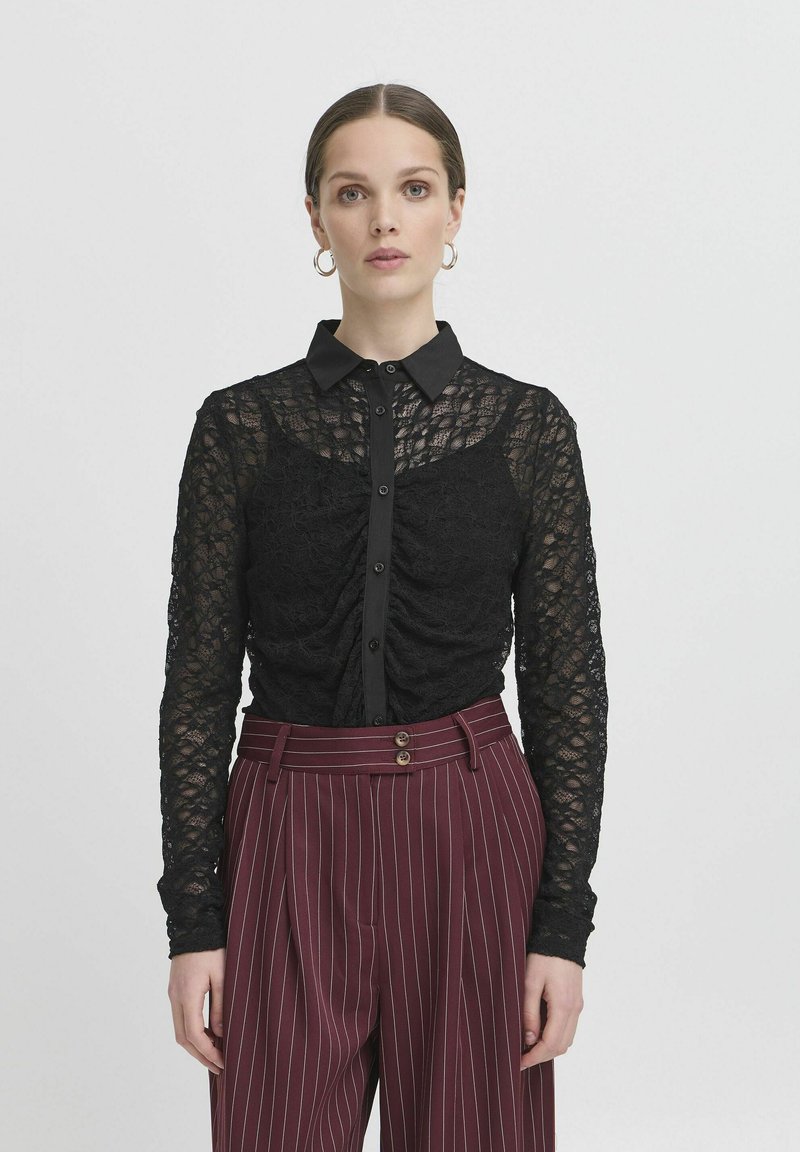Blusa in pizzo nero con chiusura a bottoni, maniche lunghe e collo; caratterizzata da dettagli arricciati sul torso, abbinata a pantaloni a righe bordeaux.