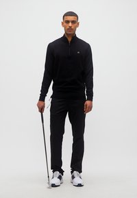 Homme portant un haut noir à manches longues et un pantalon, tenant un club de golf, debout devant un fond blanc uni.