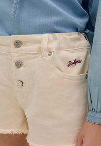 Des shorts en denim beige clair présentent un ourlet effiloché, deux boutons en argent et un logo "S" brodé en violet sur la poche avant.