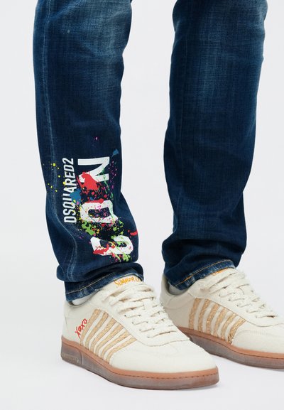 Σκούρο μπλε τζιν με πολύχρωμη σχεδίαση πιτσιλιών και λευκό λογότυπο στο κάτω μέρος του ποδιού, συνδυασμένο με λευκά αθλητικά παπούτσια με μπεζ ρίγες.