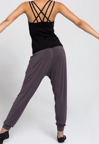 Personne portant un débardeur noir à bretelles et un pantalon gris ample, debout les bras étendus et les jambes écartées devant un fond uni.