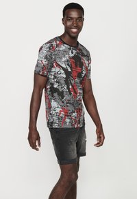 Tricou grafic de culoare gri, roșu și negru, cu modele abstracte și text. Mâneci scurte, guler rotund și croială lejeră.