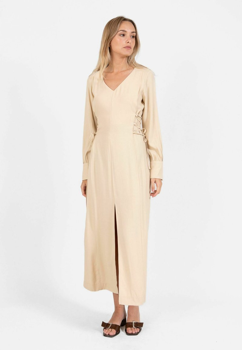 Femme debout portant une robe midi beige à manches longues, col en V, avec des liens latéraux et une fente à l'avant, associée à des sandales à talons marron.